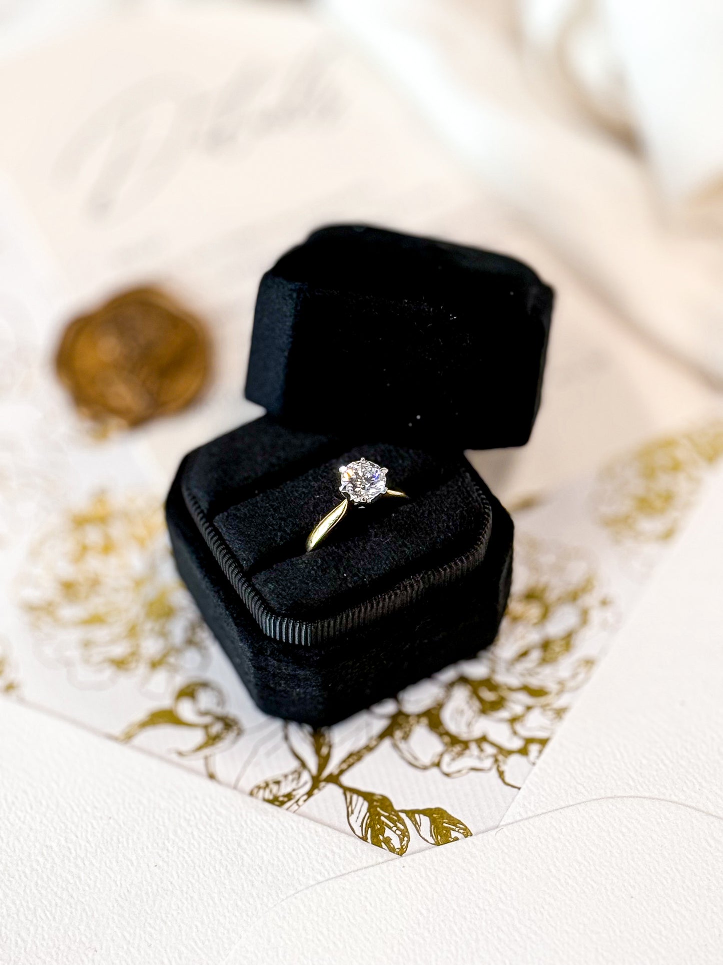Gold ring with a diamond in a black velvet box on a decorative white and gold surface.