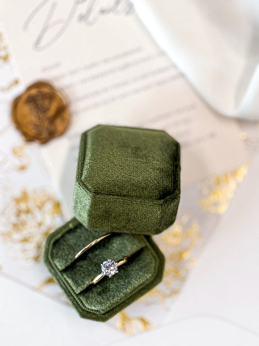Green velvet ring box with a gold ring featuring a diamond on a white surface.