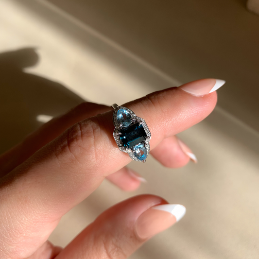 Hand wearing a ring with a blue gemstone against a neutral background